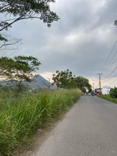 The image shows a view of a paved road in Kecamatan Margaasih, Indonesia, looking down the road towards a small town. The road is bordered by tall grass and trees on the left, and a few buildings are visible on the right. Several motorcycles are driving down the road. There are power lines overhead. In the distance, a mountain can be seen through the overcast sky.