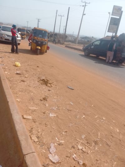 The image depicts a roadside scene in Lapai, Nigeria, under hazy daylight conditions. A two-lane paved road, dark grey in color, diagonally traverses the frame from mid-left to upper right. To the left of the road, a wide, reddish-brown dirt shoulder is visible, extensively littered with various plastic wrappers, bags, and debris, including a prominent yellow plastic bag in the foreground. A concrete curb separates this shoulder from the paved road.
On the left side of the paved road, a person wearing a red top, dark flared trousers, and a blue head covering stands next to a silver sedan, possibly a Toyota Corolla, which is partially visible. Immediately to their right, a yellow motorized tricycle, commonly known as a Keke Napep, with "NO 50" displayed on its front, is stopped on the road, facing towards the left. A red object is attached to the rear of this tricycle. Two additional yellow tricycles are visible further down the road behind the silver sedan.
On the right side of the paved road, a dark minivan or SUV is stopped. Adjacent to this vehicle, a person in light-colored traditional attire and a dark vest stands, with a smaller individual or child partially visible next to them. In the background on the right, a tall commercial sign for "NAFCON" and "QUARTZ" is present, indicative of a petrol station. Several utility poles with multiple overhead power lines are visible against the hazy sky. The background also shows residential buildings and sparse vegetation.