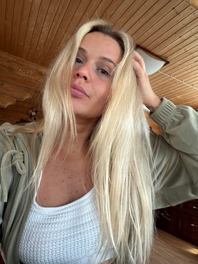 A selfie angled downwards showing a fair-skinned woman with long blonde hair. She is wearing a white knitted top and a light green jacket. Her left hand is visible near her head. The background is a wood-paneled wall and ceiling. A small window is visible on the upper left. Location cannot be determined from the image.