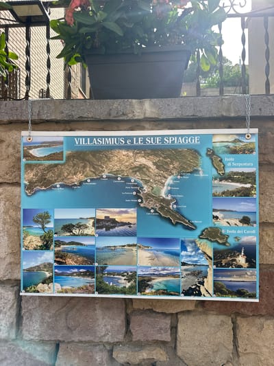 The image shows a map of Villasimius and its beaches, displayed on a stone wall. The map is bordered by smaller photographs of various beaches and islands, each numbered for reference. The title of the map is "VILLASIMIUS e LE SUE SPIAGGE." The map features coastal areas, roads, and labels for different locations such as "Porto sa Ruxi," "Cabo Boi," "Is Piscadeddus," "Campus," "Cucurreddus," "Campulongu," "Porto Turistico," "La Fortezza," "Santo Stefano," "Cala Caterina," "Cava Usai," "Cala Burroni," "Timiama," "Cala Luna," "Is Traias," and "Punta Molentis." The smaller images depict picturesque coastal scenes, including clear blue water, sandy beaches, rocky outcrops, and vegetation. On the right side of the map, two islands are highlighted: "Isola di Serpentara" and "Isola dei Cavoli," with an image of a lighthouse on the latter. The overall scene suggests a tourist information display in the town of Villasimius, Italy, likely near a coastal area. The background includes a stone wall, a metal fence, and a potted plant with flowers.
