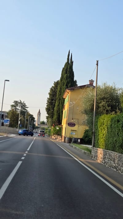 The image captures a sunlit scene on a road in Bardolino, Italy, likely in the late afternoon given the long shadows and warm light. The road, paved with asphalt, has clear white lane markings, indicating a two-lane configuration. On the right side of the road, a narrow sidewalk is bordered by a stone wall covered in vegetation. Behind the wall, a lush green hedge stands tall. A yellow building, appearing to be an osteria, is situated next to the road, featuring green shutters and a sign reading "OSTERIA CAPITEL". Two dark barrels are placed outside the building, and a tall wooden pole with wires runs alongside the property.
Dominating the right side of the frame are tall, slender cypress trees. On the left side of the road, a blue car is parked, and a white van is driving away from the camera. Further down the road, a tall bell tower, characteristic of Italian churches, rises against the clear blue sky. The overall environment suggests a semi-rural setting, typical of the outskirts of a town in Italy. The sky is a pale blue with no clouds visible.