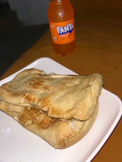 In the city of Egbe, Nigeria, a close-up, elevated view captures a meal setup on a brownish-orange surface. The primary subject is a food item, resembling a flatbread sandwich or wrap, positioned on a white, rectangular plate with rounded edges in the foreground. The flatbread exterior is light beige, with irregular brown areas from cooking. Visible through an opening in the bread is a filling consisting of irregular, light brown pieces of cooked protein, consistent with chicken or similar meat. In the background, slightly out of focus and to the right, stands a small, transparent plastic bottle of Fanta orange-flavored soda. The bottle displays condensation on its exterior and features the distinct orange and blue "FANTA" logo. The ambient lighting suggests an indoor setting. No individuals or active interactions are present in the frame.