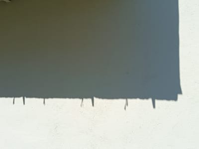 The image displays a section of a textured, white or off-white wall, likely an exterior surface, under bright sunlight. A large shadow cast by an unseen object or structure covers the upper portion of the wall, creating a stark contrast between the illuminated and shadowed areas. Along the bottom edge of the shadowed area, there are several distinct, elongated shadow shapes that suggest the presence of thin, vertical objects casting shadows onto the wall. These could be parts of a railing, fence posts, or perhaps decorative elements. The light suggests it is daytime, likely mid-morning or afternoon, given the sharp definition of the shadows. The overall impression is one of a sunny, perhaps warm day. There is no visible text in the image. The setting appears to be an outdoor architectural space, possibly in Trebinje, Bosnia and Herzegovina, as indicated by the context provided.