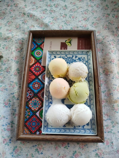 A wooden tray with a decorative insert and a blue and white patterned plate holding six scoops of ice cream. The ice cream scoops are in three rows of two. The top row has yellow and white ice cream. The middle row has light orange and light green ice cream. The bottom row has white ice cream. The tray is on a floral patterned tablecloth. The text on the insert inside the tray is in Vietnamese and mentions "Trường An, Vietnam".