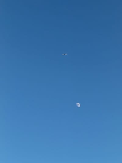 An airplane and the moon are visible in a clear blue sky, taken from Riverhead, United States of America. The airplane is white with red accents and is flying horizontally across the sky, slightly to the left of the center. The moon is in its waxing gibbous phase, with visible craters and surface details, positioned lower and to the right of the airplane.