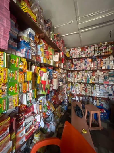 The image depicts the interior of a pharmacy in Akowonjo, Nigeria. The shelves are densely packed with a wide variety of medicines in boxes and bottles. The lighting is somewhat dim, and the overall impression is one of a busy, well-stocked establishment. In the foreground, there are two orange chairs, one of which is partially visible. A wooden stool and a black plastic bag are present on a long, narrow surface to the right. The floor is dark and appears to be tiled or concrete. The walls and ceiling are visible, showing signs of age and wear. The shelves are made of wood.