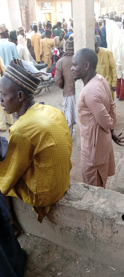 This image depicts a crowded outdoor scene in Kabuga, Nigeria. In the foreground, two adult males are prominent. On the left, a man wearing a yellow patterned tunic and a striped brown and white cap is seated or squatting on a low, rough concrete or stone barrier, facing away from the camera's direct view. To his right, another man in a light brown, long-sleeved tunic stands with his hands clasped behind his back, looking towards the right side of the frame. He exhibits a short beard and mustache.

Behind these two individuals, a dense gathering of approximately 20-30 people, primarily adult males, fills the midground and background. They are standing and moving within what appears to be a public space or market area. Many individuals wear traditional Nigerian attire, including various tunics and skullcaps. A white and red motorcycle or scooter is partially visible amidst the crowd in the midground, positioned to the left. The ground is an uneven, dusty surface of packed earth or dirt with scattered small pieces of debris. Several concrete pillars are present throughout the scene, one prominent to the left of the standing man. In the background, blurred building structures, possibly shops, are visible, one featuring a partial sign displaying the word "FRESH." The scene is illuminated by natural daylight, suggesting an outdoor environment during the day.