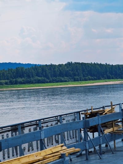 The image captures a serene river scene on a bright, partly cloudy day. The foreground is dominated by the deck of a vessel, with a weathered blue railing and a mesh fence. Stacked lumber, in various shades of yellow and brown, is piled precariously on the deck, suggesting preparation for transport or construction. The lumber is supported by a rustic wooden structure and metal poles.
Beyond the vessel's deck, a wide, calm river stretches out, reflecting the soft blues of the sky and the distant greenery. The middle ground features a sandy riverbank lined with lush green grass, giving way to a dense forest of dark green trees that blanket the rolling hills in the background. The tree line is varied, with some areas appearing more mountainous. The overall impression is one of peaceful, natural beauty.
The time of day appears to be late morning or early afternoon, given the bright sunlight casting gentle shadows and illuminating the scene. The weather is pleasant, with visible clouds scattered across a clear blue sky, suggesting a fair day. The location cues, combined with the style of the vessel and the natural landscape, are consistent with a river in a remote, possibly rural area. There is no visible text in the image. The scene evokes a sense of calm and tranquility.