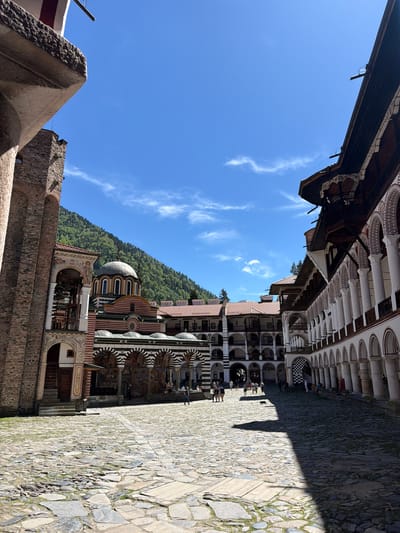 The image captures an upward view within the courtyard of Rila Monastery, located in Rilski manastir, Bulgaria. The architecture of the monastery is prominent, showcasing its distinctive design with layers of arched openings, columns, and black and white stripes. The building on the left is primarily stone and has a simple design. The courtyard is paved with cobblestones. In the background, the blue sky is visible. Groups of people are visible walking along the perimeter of the courtyard, suggesting the area is a tourist destination. A portion of the mountain covered with dense trees is behind the architecture.