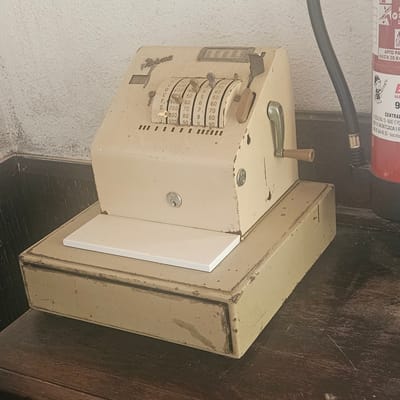 This image shows a vintage mechanical calculator, possibly a cash register or adding machine, resting on a dark wooden surface. The object is made of metal and has a cream-colored, aged finish, showing signs of wear and tear with some scratches and scuff marks. It features a series of rotatable dials with numbers and letters, likely used for calculations or to record transactions. A crank handle is attached to the top left side. A small, light-colored rectangular panel is attached to the front right side of the machine.
In the background, a textured light-colored wall is visible on the left, and a red fire extinguisher with some text in Spanish is mounted on it. The text on the fire extinguisher includes "APTO PARA HASTA 50 KV" and a logo. Additional text appears to be related to a company named "CENTRAL" with an address in Montcada i Reixac, Barcelona.
The scene appears to be indoors, possibly in a workshop, storage area, or an old-fashioned shop, given the presence of the mechanical device and the fire extinguisher. The lighting suggests it's daytime, but the exact time is not discernible. The overall impression is one of an older, possibly forgotten piece of equipment, hinting at a historical or retro aesthetic. No people are visible in the image.