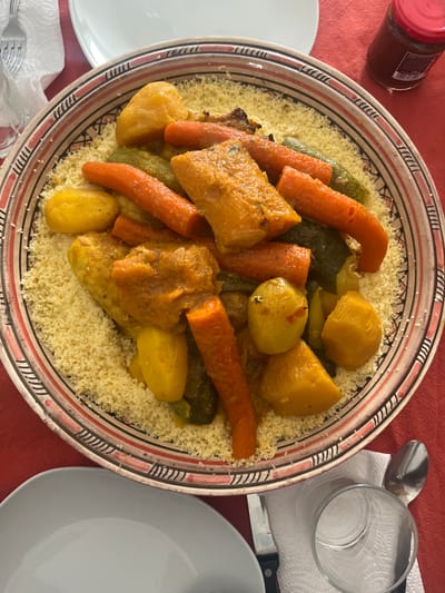 A large, round ceramic bowl, filled with couscous and a vegetable and meat stew, is centered on a red tablecloth. The couscous base is light yellow and granular. Dominating the stew are numerous elongated orange carrots, irregular yellow potato pieces, dark green zucchini or squash sections, and larger, irregular pieces of orange-yellow meat or poultry, all coated in a rich, yellowish-orange sauce. The bowl features a decorative rim with black horizontal lines, vertical dashes, and reddish-brown bands. Surrounding the bowl are various dining items: a white dinner plate partially visible in the upper left, another white dinner plate in the lower left, a clear drinking glass with a subtle pattern in the upper left, a small glass jar with a red lid (label showing "200 g") in the upper right, and a clear drinking glass, a silver spoon, and a knife handle resting on a white paper napkin in the lower right. The scene depicts a meal prepared in Le Puy-en-Velay, France.