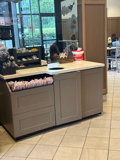 The scene shows an interior location in the city of Stone Mountain, United States of America. The location has large windows with "PLAY" printed on the left side of the window. The window has an unobstructed view of the exterior, revealing green trees. The interior includes a counter with a sanitation station, including a container of wipes. The counter also includes an opening, and below this opening, there is a holder with straws. The location is a fast-food restaurant where patrons can get condiments such as ketchup and sauce. The floor is tiled. There are no interactions taking place.