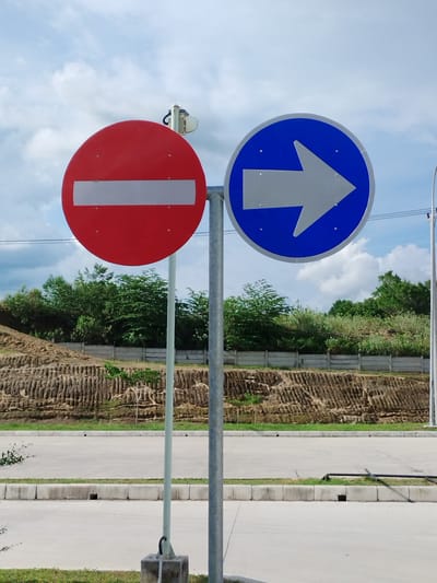 In Keureuweungkrueng, Indonesia, a traffic signpost stands prominently in an outdoor setting. The signpost supports two circular signs: a red sign with a white horizontal bar, indicating "Do Not Enter," and a blue sign with a white arrow pointing to the right, signifying a mandatory direction. The signs are mounted on a sturdy metal pole, which is anchored to the ground by a concrete base. Behind the signs, a gently sloping embankment is visible, covered with sparse vegetation and featuring a distinct, terraced pattern of soil erosion. A concrete wall runs along the top of the embankment, and beyond that, a line of trees forms a dense green backdrop. In the foreground, a paved road stretches across the image, bordered by a strip of grass and concrete barriers. The sky above is a mix of blue and white clouds, suggesting fair weather. No people or animals are present in the scene.