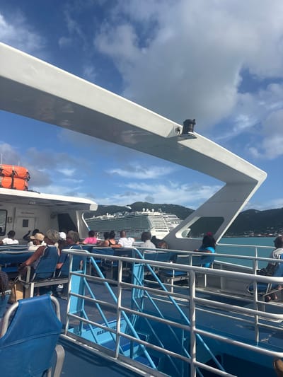 The image displays a view from the upper deck of a passenger ferry or catamaran in Road Town Harbor, British Virgin Islands. The vessel's white superstructure forms an angular overhead beam and a bulkhead on the left. A black mounted device, possibly a light or sensor, is visible on the overhead beam. Below, a blue deck area is fitted with multiple rows of blue seats with white frames. Approximately 15 to 20 individuals, mostly adults, are seated as passengers, facing generally towards the right or away from the camera. Visible elements on the ferry include an orange life raft container on the left, a "NO SMOKING" sign on a white wall, and a partially visible sign indicating "MAXIMUM PASSENGERS 81" on a blue railing. In the midground, the blue-green water of the harbor is visible, with a large, white, multi-deck cruise ship prominent towards the center-right. The cruise ship is docked or anchored close to a landmass. The background consists of green, hilly terrain consistent with the islands of the British Virgin Islands, under a blue sky with scattered white cumulus clouds. Passengers are observed in a passive state, seated and observing the environment; no specific active interactions are depicted.