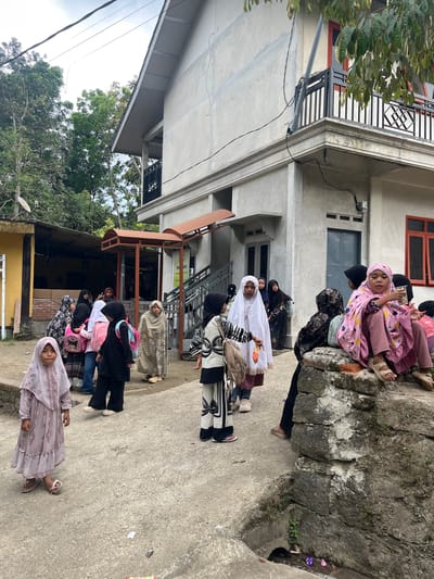 The image captures a group of children, mostly girls, gathered outside a building in Jurit Baru, Indonesia. The building, a simple concrete structure with an orange-framed window visible, appears to be either a school or a community center. Some of the children are wearing traditional Islamic attire, including hijabs and modest dresses. A few of the children are carrying backpacks, suggesting they may be returning from or going to school.
The children are positioned on a paved area, with a low stone wall to the right of the frame. The environment appears somewhat rural, with trees and foliage visible in the background. The sky is overcast, indicating a cloudy day.
Several of the children are interacting with each other, with some looking towards the camera. One girl in the foreground, wearing a light pink hijab and dress, is looking directly at the camera. Another girl, seated on the stone wall, is holding a small container and appears to be engaged in conversation with someone.
The overall scene suggests a moment of everyday life for these children in their community.