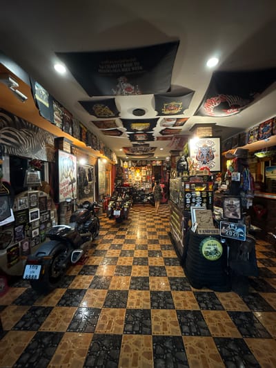 The image depicts the interior of a motorcycle shop in Kalai, Thailand. The shop is filled with numerous motorcycles, displayed both inside and possibly in an adjacent open area. The walls and ceiling are adorned with a multitude of posters and banners, many of which appear to be related to motorcycle events or clubs, featuring graphics, text, and images. The floor is tiled in a checkered pattern of brown and black. In the foreground, a black motorcycle with a license plate reading "100 8888" is prominently displayed. Further into the shop, more motorcycles are visible, including one with a license plate reading "2025". A woman in a checkered dress is walking away from the viewer towards the back of the shop. The overall atmosphere suggests a dedicated space for motorcycle enthusiasts, showcasing merchandise and possibly offering services. The shop appears to be well-lit, with artificial lighting from the ceiling.