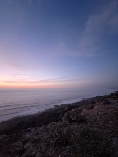 The image depicts a twilight scene over the Atlantic Ocean, viewed from a rocky cliffside. The sky displays a gradient of colors, transitioning from deep blue at the top to soft purples and pinks near the horizon. A thin band of orange light is visible at the very edge of the horizon where the sky meets the ocean. The ocean itself is calm, with faint, elongated waves reflecting the muted sky colors. A thin layer of fog or mist hovers over the water's surface, obscuring the details of the distant sea.

The foreground is dominated by a rugged, uneven terrain covered in rocks, pebbles, and sparse vegetation. The rocks are dark in color and appear to be part of a natural cliff formation. Some patches of green grass and small plants are visible amongst the rocks, especially in the lower right portion of the image.

The location is identified as a cliffside in the city of Ahrara, Morocco. The overall impression is one of a serene and natural coastal landscape at dusk. There are no people, animals, or man-made structures visible in the image. The scene is static and quiet, emphasizing the natural beauty of the coastal environment.