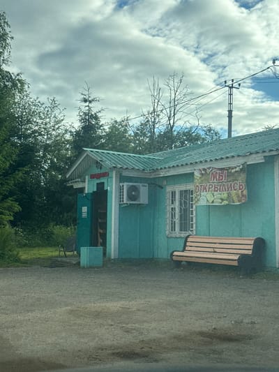 A slightly tilted, medium shot captures a small, turquoise-colored building with a corrugated metal roof. The building appears to be a shop or kiosk. A banner with text in Cyrillic script, reading "МЫ ОТКРЫЛИСЬ" (We are open), is affixed to the right side of the building, above a window with a metal grate. The banner also features an illustration of produce, such as apples and possibly cabbage. An air conditioning unit is mounted on the wall to the left of the window. To the right of the building, a wooden park bench with black metal supports sits on the ground. To the left of the building, an open doorway reveals a dark interior with some shelving and what appears to be a ladder. A light blue rectangular object sits in front of the doorway. The building is situated on a gravelly or dirt surface, with a small patch of grass and some trees visible in the background. Power lines and a utility pole are visible against a cloudy sky. The scene is set in a somewhat rural or suburban area, likely on the outskirts of Zhilgorodok, Russia. There are no people visible in the image. The overall impression is one of a newly opened small business in a modest setting.