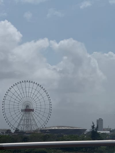 An exterior, eye-level daytime shot captures a panoramic view of the Sun World Da Nang Wonders complex and surrounding urban landscape in Ngũ Hành Sơn Ward, Da Nang, Vietnam, under a partly cloudy sky. The dominant feature is a large, white Ferris wheel, identified by "SUNWORLD" in red text on its central hub. The wheel exhibits a complex radial spoke structure and approximately 64 passenger gondolas. To the right of the Ferris wheel's base, a large, dark-colored, elliptical, dome-shaped building is prominent. Smaller structures with traditional Vietnamese architectural elements, including tiered, curved roofs, are partially visible at the base of the Ferris wheel, obscured by green foliage. In the far right background, a tall, light-colored, multi-story rectangular building is visible against the horizon. The immediate foreground features a silver-colored metal railing, consisting of two horizontal bars. A dense band of green trees and shrubs occupies the mid-ground, separating the foreground railing from the major structures. The sky is largely covered by a mix of light and dark grey cumulus clouds, with some patches of lighter blue visible in the upper portion of the frame. No individuals, specific actions, or interactions are discernible within the scene.