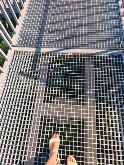 The image shows a person's feet wearing tan sandals standing on a metal grate walkway. The walkway is elevated, with grass and foliage visible beneath the grate. The sunlight casts shadows across the walkway, suggesting it is daytime. The image is taken from a top-down perspective, focusing on the walkway and the person's feet. The setting appears to be outdoors, possibly in a park or recreational area, and could be located in the city of Akhmerovo, Russia, as specified. There are no other individuals visible in the frame.