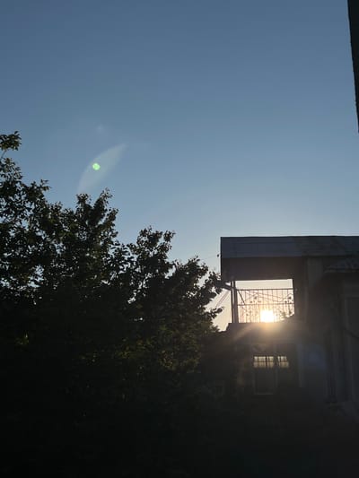 A vertical, medium-shot photograph captures a clear blue sky with a faint green lens flare near the top left. In the foreground, a silhouette of dark green foliage from a tree occupies the left side of the frame. To the right, the upper portion of a building is visible, featuring a dark-colored, angled roof. Below the roof, an open balcony or porch area is outlined, with a decorative metal railing. Through this opening, the sun is visible, emitting bright rays and creating a flare effect. Beneath the balcony, a window with multiple panes reflects the sunlight. The lower part of the building and its surroundings are obscured by deep shadows. The scene is set in the city of Ilavche, Ukraine.