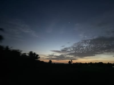 The image captures a twilight sky over a silhouetted landscape in Earipalayam, India. The sky is a gradient of deep blues at the top, transitioning through lighter blues and purples, to a soft orange and yellow hue near the horizon, indicating dusk or dawn. Wispy clouds are scattered across the sky, with a particularly dense and textured formation visible in the upper right portion. A crescent moon is faintly visible in the upper sky.

The foreground is dominated by dark silhouettes of palm trees and other vegetation, suggesting a tropical or sub-tropical environment. The blurry motion of the foliage on the left side implies either movement of the camera or wind. A low-lying landmass with more trees is visible along the horizon. The overall mood is serene and peaceful, evoking the quiet transition between day and night. There is no visible text in the image.