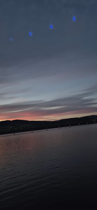 The image depicts a serene sunset scene over water, likely a fjord or sea, near Biri, Norway. The sky is painted with soft hues of pink, orange, and gray, indicating twilight. Wispy clouds stretch across the sky, reflecting the fading sunlight.

A prominent feature in the image is a long pier or bridge extending over the water. It is constructed with vertical wooden supports and a horizontal walkway, disappearing into the distance. The water's surface is calm, with subtle ripples reflecting the colors of the sky.

Along the far bank, a dark silhouette of a hilly landscape is visible, dotted with faint lights that suggest a settlement or scattered houses. The overall mood is peaceful and tranquil, characteristic of a quiet evening in a natural setting. The blue, star-like shapes scattered in the sky are likely artifacts from the camera or editing process and not part of the natural scene. No visible text is present in the image.