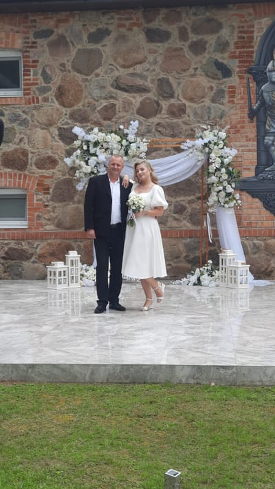 A man and a woman are standing next to each other in front of a stone building. The man is wearing a black suit and a white shirt, and the woman is wearing a white dress. They are both smiling. There is a wedding arch behind them, decorated with white flowers and draped with white fabric. In front of them, there are two white lanterns. The ground is paved with light-colored tiles. To the left of the couple, there is a window in the stone wall. To the right, there is a mural of a knight in armor. The background is a stone wall with a brick facade on the left. The overall setting appears to be an outdoor wedding ceremony. The location is identified as Жодзішкаўскі сельскі Савет, Belarus.