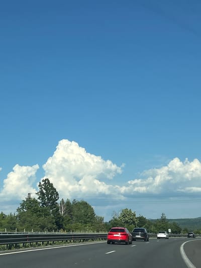 The image depicts a highway scene with a clear blue sky and scattered white clouds. In the foreground, several cars are traveling on a multi-lane asphalt road, with a guardrail separating the lanes from the shoulder. A red SUV is in the left lane, followed by a black SUV in the right lane, and a white sedan further ahead. Beyond the guardrail, there is a line of lush green trees and vegetation. In the distance, rolling hills covered in trees are visible. The scene appears to be set in a rural or suburban area, possibly on the outskirts of Novi han, Bulgaria, given the presence of a highway and the natural landscape. The lighting suggests it is daytime, likely late morning or afternoon. There are no people visible in the image.