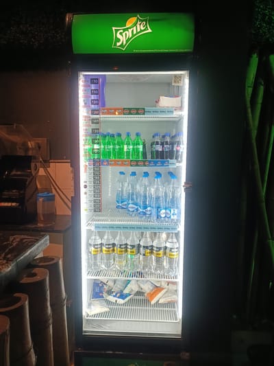The image shows an upright, illuminated cooler stocked with various beverages, including Sprite, Fanta, and Kinley water. The cooler is located in what appears to be a small shop or convenience store, with a counter and a cash register visible to the left. Bamboo-like pillars are present in the foreground, suggesting an outdoor or rustic setting within a covered area. The lighting is dim, indicating it is likely evening or nighttime.
The cooler's shelves are filled with plastic bottles and cans of soft drinks and water. Price labels are visible on the cooler door, indicating prices in Indian Rupees. Some labels mention "PET" or "Can" along with volume measurements. Brands like Sprite, Fanta, and Kinley are prominent. At the bottom of the cooler, there are some packets of what appears to be curd.
A small QR code is visible on the inside of the cooler door, likely for payment or promotional purposes. The overall scene depicts a retail environment where cold beverages are sold, with a focus on the products displayed in the cooler. There are no people visible in the image. The setting seems to be a warm climate, given the type of beverages and the apparent outdoor elements.
