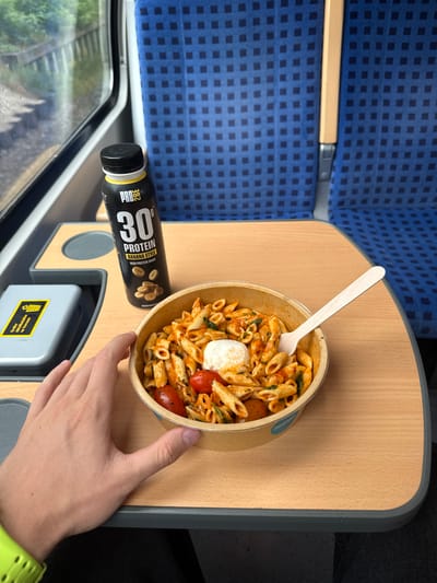 In a train carriage with blue patterned seating, a person's left hand, wearing a bright green watch, rests on a wooden tray table. On the table are a black bottle of "PRO 360 PROTEIN BANANA STYLE" and a bowl of pasta with tomatoes and white cheese, served with a wooden fork. A grey plastic tray with a yellow sticker is also on the table. The window shows a blurred view of greenery and tracks, suggesting travel outside of Wittenberg, Germany.