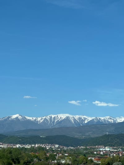 The image shows a clear blue sky with some wispy clouds. On the right side of the image is a mountain range with snow-capped peaks. At the base of the mountains is a town, likely Moshtanets, Bulgaria, with buildings and trees. The image is rotated 90 degrees counter-clockwise.