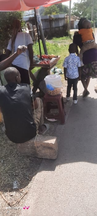 The image depicts a roadside market scene in Homa Bay, Kenya. In the foreground left, an adult male with short hair, dressed in a black long-sleeved shirt and light-colored striped pants, sits on a large concrete block. He wears light-colored flip-flops, and a thin, light-colored string extends from his hand. To his right, positioned on the asphalt pavement, a small, reddish-brown plastic stool supports a clear plastic bucket filled with light-brown spherical items, likely roasted groundnuts.
Adjacent to the seated male and stool, a makeshift market stall with a red canopy overhead displays various produce, including red tomatoes and other yellow/orange fruits, some contained in a bag. An adult female with braided hair, wearing a white short-sleeved top, stands behind this stall, appearing to attend it. To the right of the stall, an adult female in a dark top and patterned skirt carries a small child on her back; the child wears an orange t-shirt and light-colored pants. A young boy, wearing a blue and white patterned short-sleeved shirt and dark pants, stands near this female, facing the market stall.
The environment includes an asphalt road on the right, transitioning to a dry, grassy, and dirt area on the left with scattered debris. The background shows foliage, a corrugated metal fence, and distant structures under bright daylight, casting clear shadows.