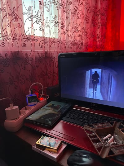 A slightly rotated, eye-level shot shows a desk surface cluttered with objects. A laptop with a Dell Inspiron label displays a figure walking through a dimly lit tunnel. Next to it is a smartphone showing an image of a person with a tattoo. An ashtray filled with cigarette butts sits on the laptop's keyboard area. A power strip with a charging block labeled "TUZ" is visible. A decorative lighter is present near the edge of the desk. The background features a window covered with patterned curtains. The location is Charentsavan, Armenia. There is no human presence beyond the images displayed on the screens.