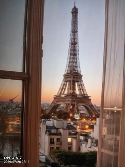 The image shows a view of the Eiffel Tower in Paris, France, seen through a window. The Eiffel Tower is centrally located and dominates the skyline. The scene is set at dusk or dawn, as indicated by the soft, warm glow of the sky and the illuminated cityscape below. The foreground features a window frame and a sheer curtain on the right side, suggesting an indoor perspective. On the left side, a portion of another window pane is visible, reflecting a hint of the cityscape and the sky. The city below the Eiffel Tower is densely built with buildings, some of which are illuminated with lights. The overall impression is one of a romantic or picturesque view of Paris.

There is no indication of the city of Құмөзек, Kazakhstan, in this image. The content of the image is entirely focused on Paris.