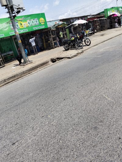 This image presents a street scene in Mkomani ward, Kenya, captured during daylight. The foreground features an asphalt road, transitioning to an elevated concrete sidewalk on the right. The sidewalk exhibits a significant depression or pothole towards the center. A row of single-story commercial structures lines the right side of the street.
The leftmost building is a shop with a green facade, displaying white text including "FOODIE NATION," "TEA ROOM SHOP," and "DAVIS MILK & MORE." A grey utility pole stands on the sidewalk in front of this shop. A person in dark attire is seated or squatting next to the utility pole, facing right. Another male individual, wearing a white shirt and dark pants, stands with arms elevated near the entrance of the green shop.
Two motorcycles are parked on the sidewalk further down the street; the closer one is dark-colored with yellow accents. Beyond these, a series of smaller shops or stalls continues, some shaded by awnings, including a white-and-red striped umbrella and a pink umbrella further in the distance. Indistinct figures are visible near these distant shops. Various merchandise is displayed outside the shops, often in crates or on shelves. The sky is light blue with scattered white clouds.