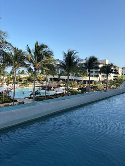 The image depicts a view of a resort pool area in Punta Sam, Mexico, on a clear day. In the foreground, a large infinity pool occupies the bottom half of the frame, with ripples on its blue surface. A concrete edge separates the pool from the background. Beyond this edge is a resort area with a large, light-colored building in the center, featuring multiple stories and balconies. Several palm trees are scattered throughout the scene, with lush green foliage. A swimming pool, occupied by several people swimming and relaxing, is visible in the middle ground. Lounge chairs and umbrellas are set up around the pool. The sky is a clear, bright blue.