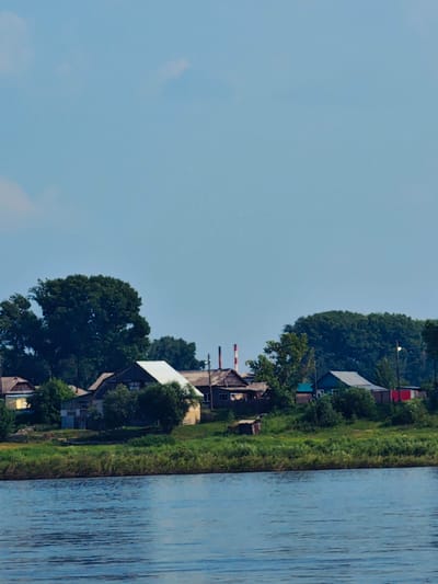 The image depicts a pastoral scene along a riverbank in Yeniseysk, Russia. In the foreground, the calm, blue water of the river reflects the sky. The middle ground features a verdant, grassy embankment lined with trees. Beyond the embankment, a cluster of residential buildings is visible, characterized by their rustic architecture, including pitched roofs and wooden structures. Two prominent smokestacks with red and white banding rise from one of the buildings, suggesting industrial activity. The sky above is a clear, pale blue with faint clouds. The overall impression is one of a rural settlement bordering a waterway, with a hint of industry present.
