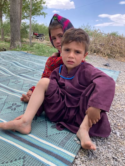 Two children are sitting on a striped mat outdoors. One child, with short brown hair and light skin, is wearing a long-sleeved, dark purple robe-like garment and a blue beaded necklace. They are looking directly at the camera. The other child, with a patterned red and black headscarf, is looking from behind the first child. A motorcycle can be seen in the background, along with trees, gravel, and a cloudy sky. The location is Qilla Saifullah, Pakistan.