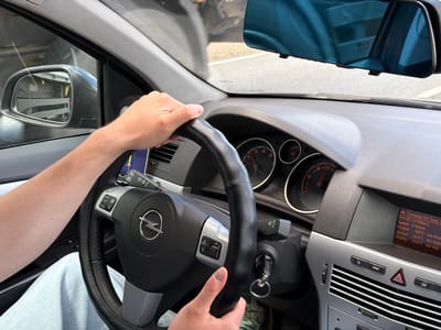 The image shows a close-up of a person's hands on a steering wheel of a car. The person is wearing a light blue shirt. The steering wheel is black and has the Opel logo in the center. The dashboard of the car is visible, including the speedometer and other gauges. The car appears to be in motion, as suggested by the reflection in the rearview mirror. The environment outside the car is blurred, but it seems to be a sunny day. There is no information in the image to indicate the location as Alnashi, Russia.