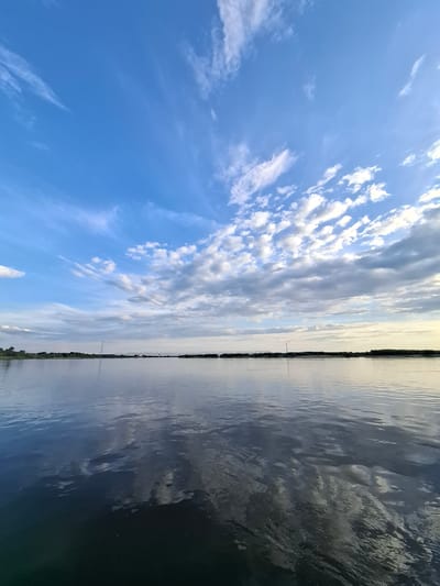 The image shows a wide, calm river with a distant tree line and a bridge visible across the water. The sky is a vibrant blue with scattered white clouds, some of which are reflecting on the surface of the water. The lighting suggests it is either early morning or late afternoon. The scene is peaceful and natural, with no people or specific structures of interest apart from the bridge. The location is identified as Vvedenovka, Russia.