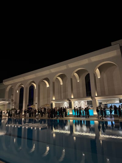 An outdoor night event is taking place in front of a modern building with arched entrances in Tsilkani, Georgia. A large, dark blue swimming pool dominates the foreground, its surface reflecting the illuminated building and the lights from the event. Floating in the pool are several white, rectangular structures that appear to spell out "GOLD LEAD". A crowd of people, dressed in formal attire, are gathered along the edge of the building, some standing by bars, others conversing. The building itself is light-colored and features a repeating pattern of large arches. The sky is dark, indicating it is nighttime. There are also some yellow umbrellas visible among the crowd.
