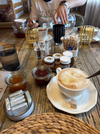 In La Ila - Stern - La Villa, Italy, a table is set for breakfast or brunch. A person's hands, with colorful nail polish, are visible holding a small glass bottle over a cup of coffee. The table is made of wood and features a variety of breakfast items, including a glass teapot with red liquid, several decorative glasses, small jars of jam and what appears to be Nutella, a white bowl filled with individually wrapped pats of butter, and a coffee cup with a frothy beverage and a spoon. There are also packets of sugar and creamer. The scene is captured from a top-down perspective, focusing on the arrangement of the food and drinks.
