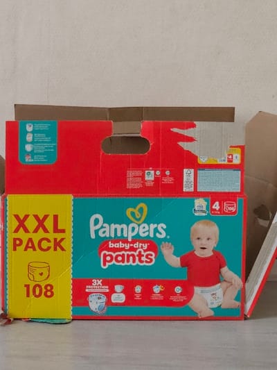 The scene, located in Villeneuve-Saint-Georges, France, depicts a large, open cardboard box for "Pampers baby-dry pants" diapers, positioned indoors against a plain, light-colored wall and resting on a light-colored floor. The box prominently displays "Pampers. baby-dry pants" branding, featuring a red upper panel and a teal lower panel. A yellow vertical strip on the left indicates "XXL PACK" and "108" diapers. The front panel includes an image of a Caucasian baby with blonde hair, wearing a red t-shirt and a diaper, smiling and sitting with one hand raised. Additional text on the box specifies "3X PROTECTION," size "4" for "9-15 kg," and a quantity of "108" diapers. Various small icons and French language text providing product details and company information (e.g., "P&G FRANCE SAS") are also present. The top flaps of the cardboard box are open and show signs of minor tearing. A partial view of another cardboard element is visible on the right edge of the frame. The environment is consistent with an interior space under ambient lighting.