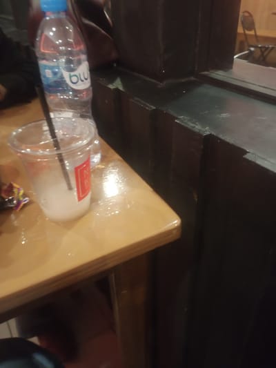 A clear plastic cup with a straw and a milky white beverage is positioned on a wooden table. Next to it, a partially visible plastic water bottle with a blue cap and a white label bearing the text "blu" is also on the table. The table surface appears wet and reflective. In the background, a dark wooden structure with a reflective surface suggests a counter or a wall with a mirror. A chair with a metal frame and wooden seat is visible in the reflected area. The overall scene suggests an indoor setting, possibly a cafe or restaurant. This image was taken in Pontianak, Indonesia.