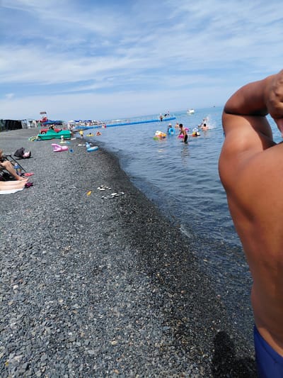 The image depicts a daytime scene at a pebble beach with the sea in the background. In the foreground, a person's arm and shoulder are visible, obscuring a portion of the frame. The beach is covered in dark grey pebbles, with several pairs of sandals and a single sandal placed on the pebbles. Further back, there are several beach umbrellas and tents, suggesting a populated area. Children and adults are in the water, some with inflatable rings. A small red and green pedal boat is visible on the shore. The sea is calm, and the sky is blue with scattered white clouds. The location is identified as a beach in Ақши, Kazakhstan.
