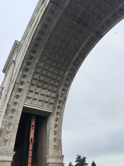The image shows a close-up, low-angle view of a large, arched stone structure with intricate decorative patterns on its underside and sides. The arch has a coffered ceiling, with square recessed panels, and a border of circular motifs. The structure appears to be made of weathered stone, with some cracks visible in the ceiling. Beneath the arch, an opening reveals a dark interior with a large, reddish-brown industrial mechanism. In the distance, silhouetted against a cloudy sky, are the tops of several coniferous trees. Three small birds are visible flying in the sky. The overall impression is of an imposing, possibly historical, piece of architecture, likely part of a larger structure such as a bridge or a gate. The location is identified as Vasil'ki, Russia, suggesting this is a landmark within that city.