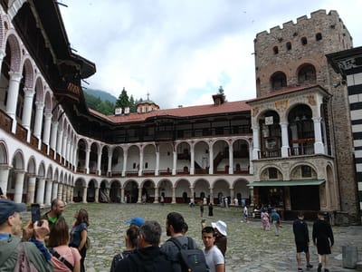 This media file depicts the courtyard of the Rila Monastery in Bulgaria. Numerous people are present, appearing to be tourists exploring the historic site. The architecture of the monastery is a prominent feature, with its white colonnades, arched walkways, and red-tiled roofs creating a visually striking scene. A tall, stone tower, a significant element of the monastery's defensive structure, stands prominently in the background.
The sky is overcast with clouds, suggesting a cool or temperate weather condition. The time of day appears to be daytime, given the ambient light. The ground of the courtyard is paved with cobblestones. The people are casually dressed, indicating a leisurely visit. Some individuals are taking photographs, capturing the essence of the location. The overall atmosphere seems calm and observant, with people engaged in sightseeing and appreciating the historical and architectural significance of the Rila Monastery. The scene is set in a mountainous region, hinted at by the treeline and mist-covered hills in the distance. No visible text is present in the image.