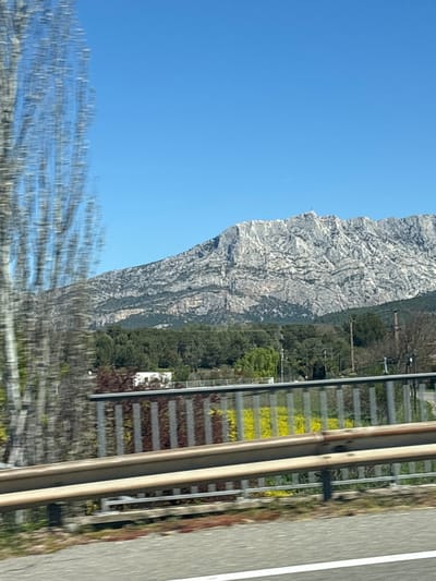 The image shows a view from a moving vehicle, likely a train or car, in Meyreuil, France. A large, rocky mountain occupies the left side of the frame, contrasting against a clear blue sky. A blurred foreground suggests movement, possibly foliage along the roadside. To the right, a series of parallel lines indicate a roadside barrier. Beyond the barrier, there's a railway line with brown and yellow vegetation. The edge of a road is visible on the far right. The perspective is slightly skewed due to the angle of the shot from a moving vehicle.