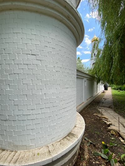The image depicts a low-angle, sideways view of a white brick wall curving into the foreground, with a pathway receding into the distance on the right. Overhanging green foliage from a tree creates a partial canopy in the upper left portion of the frame. In the background, a white church tower with a golden dome and a spire is visible against a blue sky dotted with white clouds.
The pathway, made of light-colored rectangular stones, is bordered by dark soil and scattered rocks, with some patches of dry grass and low-lying green plants, including a prickly pear cactus with a small yellow flower. The white brick wall appears to be part of a building or structure with rounded architectural elements, showing a repeating pattern of bricks. The overall scene suggests an outdoor setting, possibly a courtyard or garden area adjacent to a religious building.
The lighting indicates it is daytime, with sunlight casting shadows and highlighting textures. The weather appears fair with a clear blue sky. No people are visible in the frame. The visual elements suggest a peaceful and serene atmosphere, typical of a quiet religious or historical site. The location context provided is Plastunovskaya, Russia.